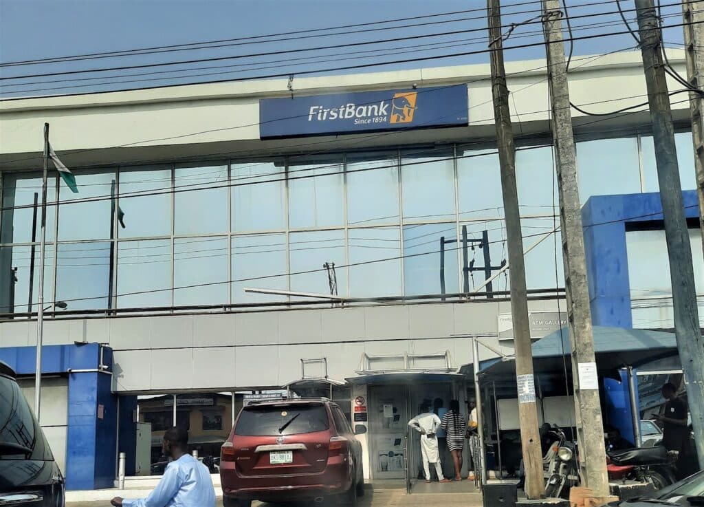 The picture shows a branch of First Bank in Ikeja, Lagos. The facade has a lot of glass windows. There is an ATM gallery on the ground floor that is accessible from the road. A ramp is provided for wheelchair users. A red SUV is parked outside the building and about four people are under the ATM shed.