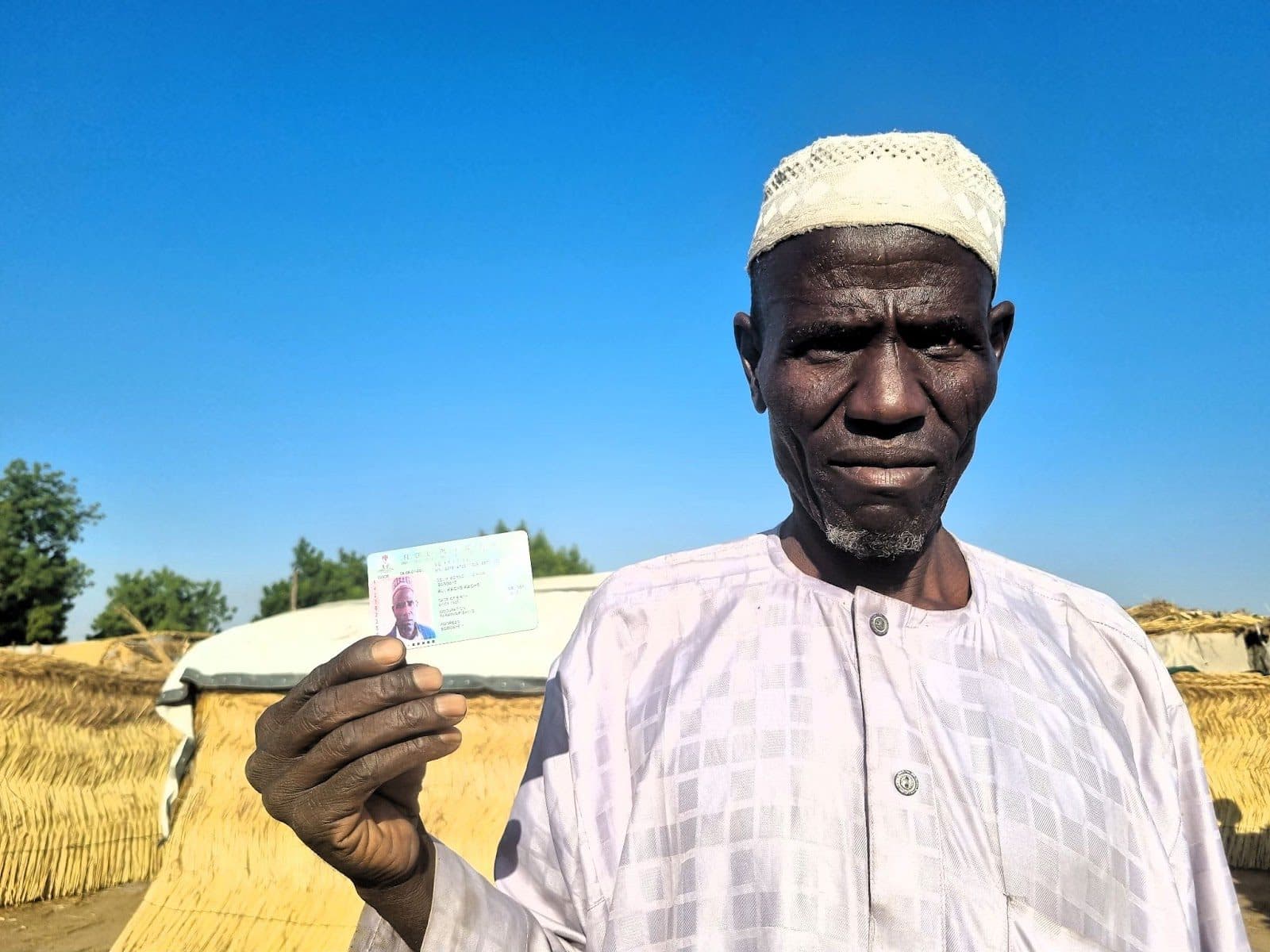 Man in traditional attire holding an ID card with thatched huts in the background under blue sky.