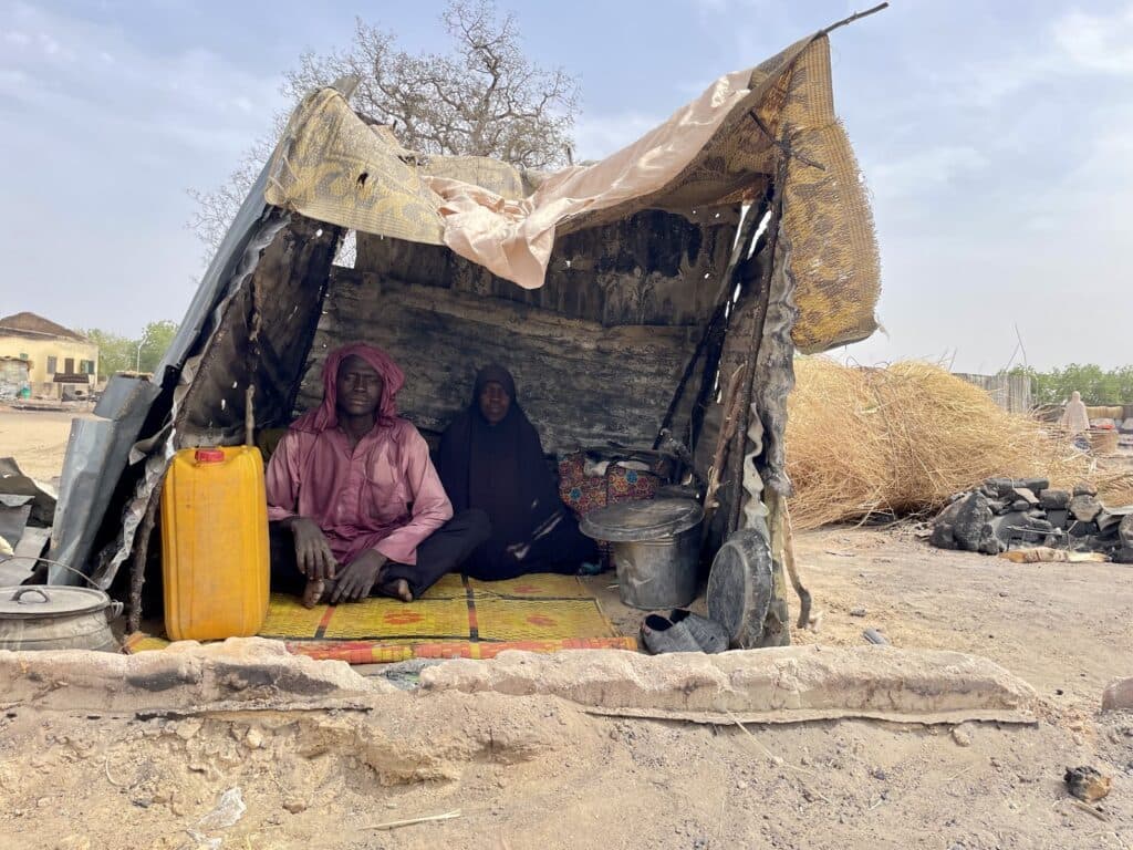 Two people sitting inside a makeshift shelter with a yellow container and cooking pots, in a rural setting.
