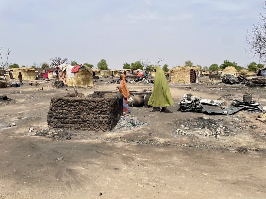 Two individuals walking among the remains of burnt huts in a devastated village.