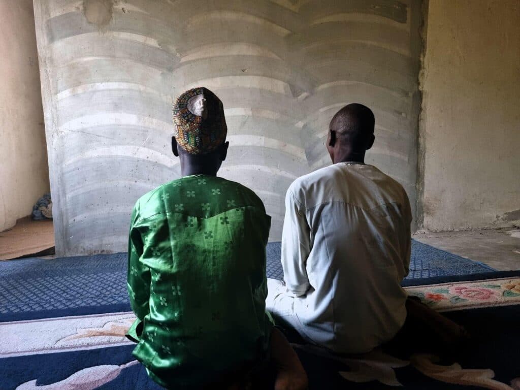 Two people sitting side by side against a curved concrete wall, one in a green shirt and the other in white.