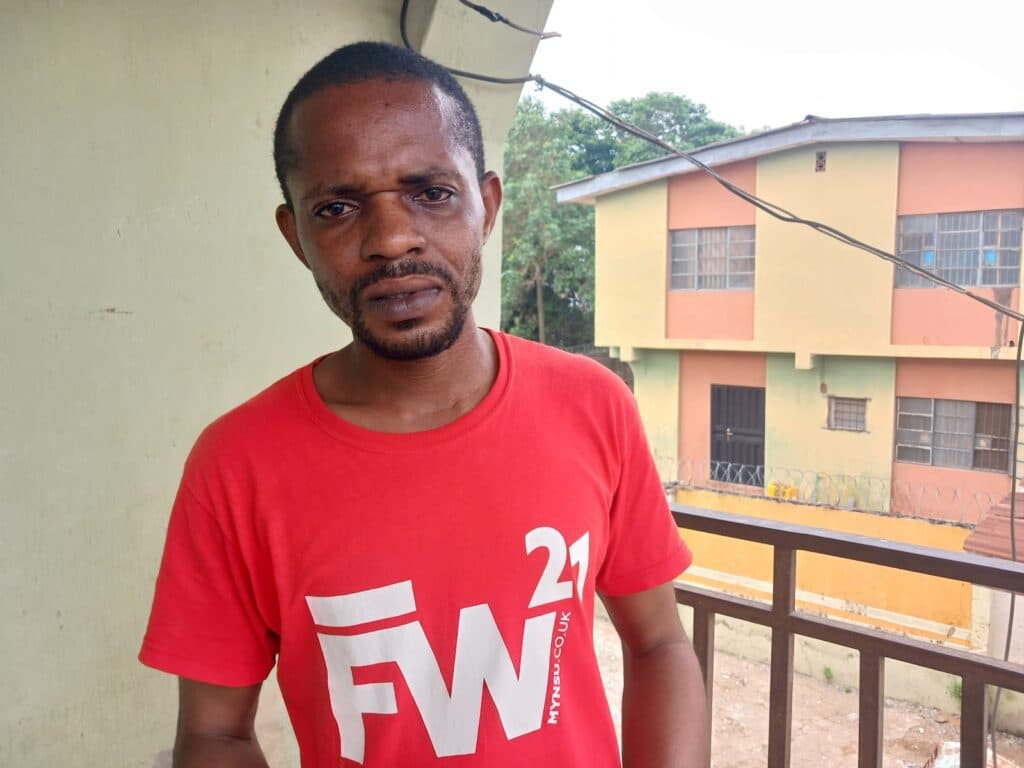 Man in red shirt standing on balcony with a two-story building in the background.