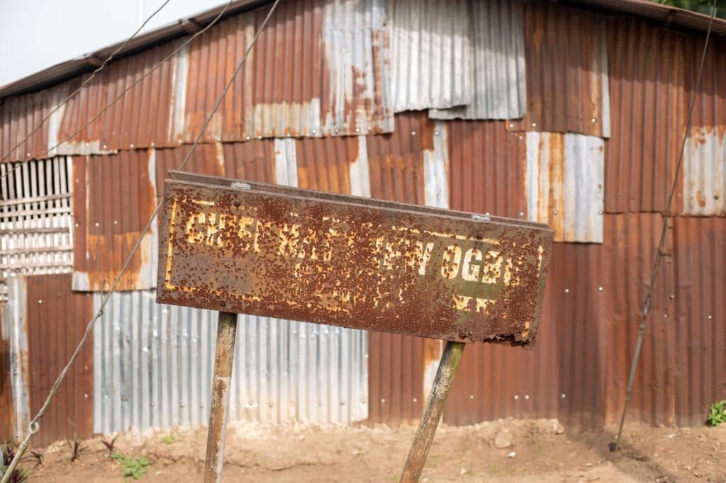 Rusty metal sign with indistinct lettering in front of a corrugated iron wall.
