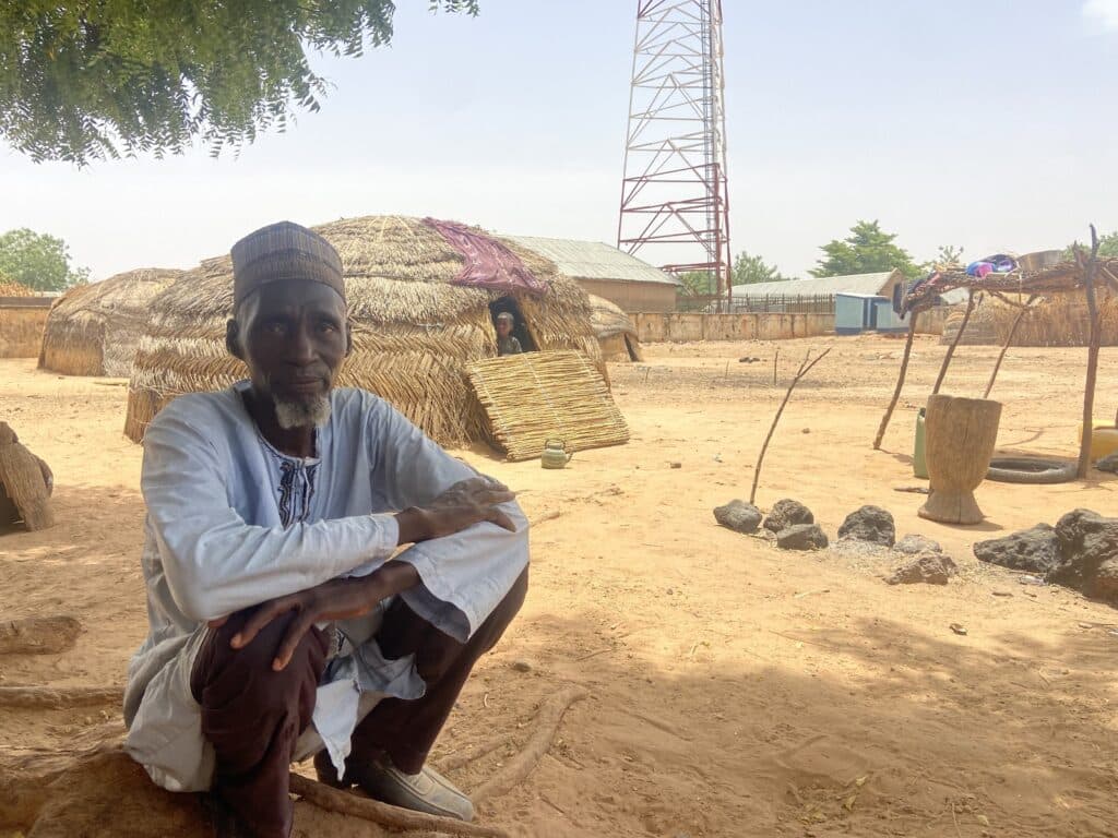 Man in traditional clothing sits in front of thatched huts with a communication tower in the background.