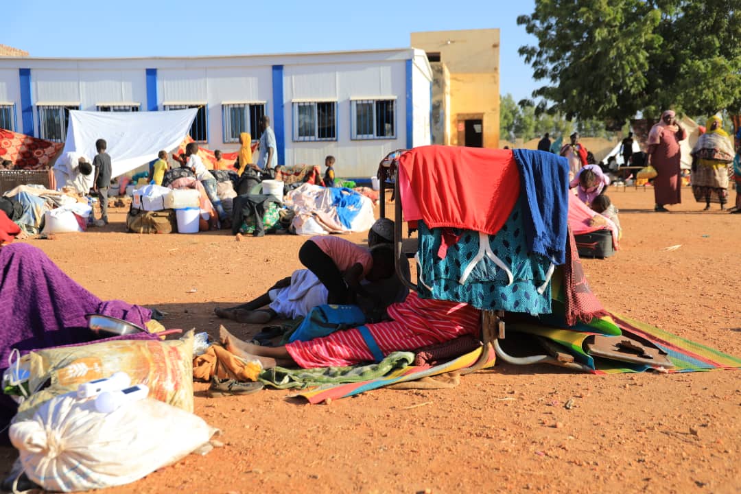 People with belongings at a temporary outdoor setup, possibly a camp, with shelters and a clear sky.