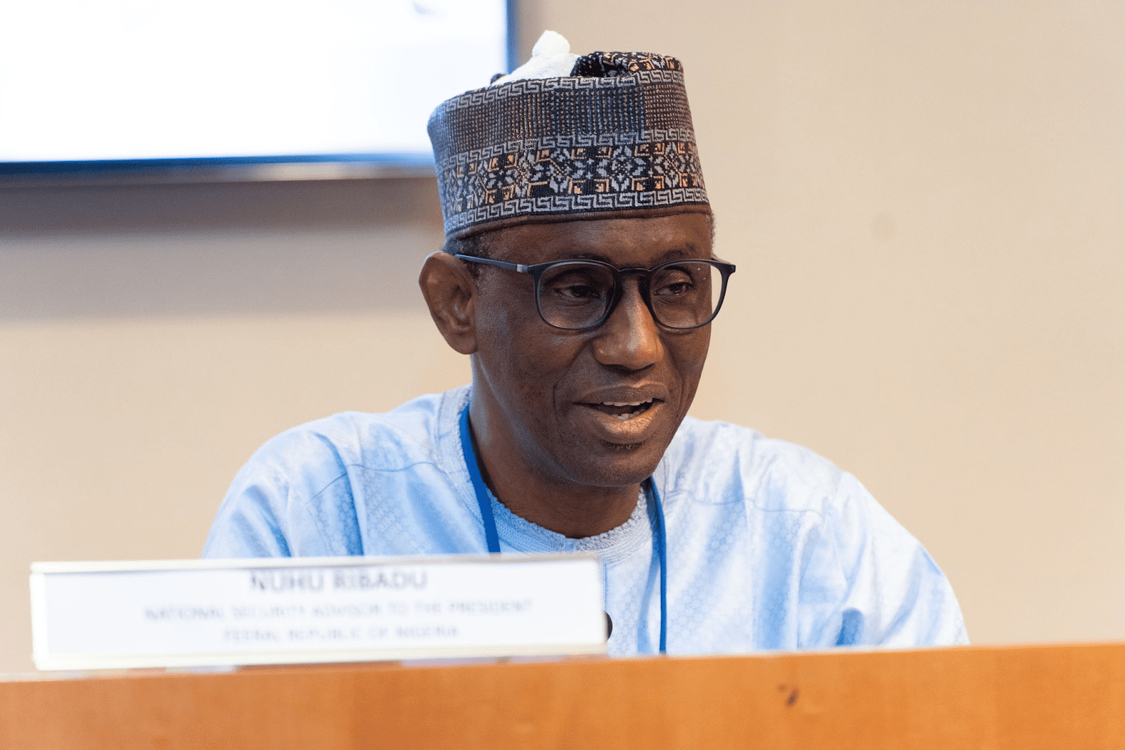 Man in traditional attire with glasses sitting behind a nameplate at a conference.