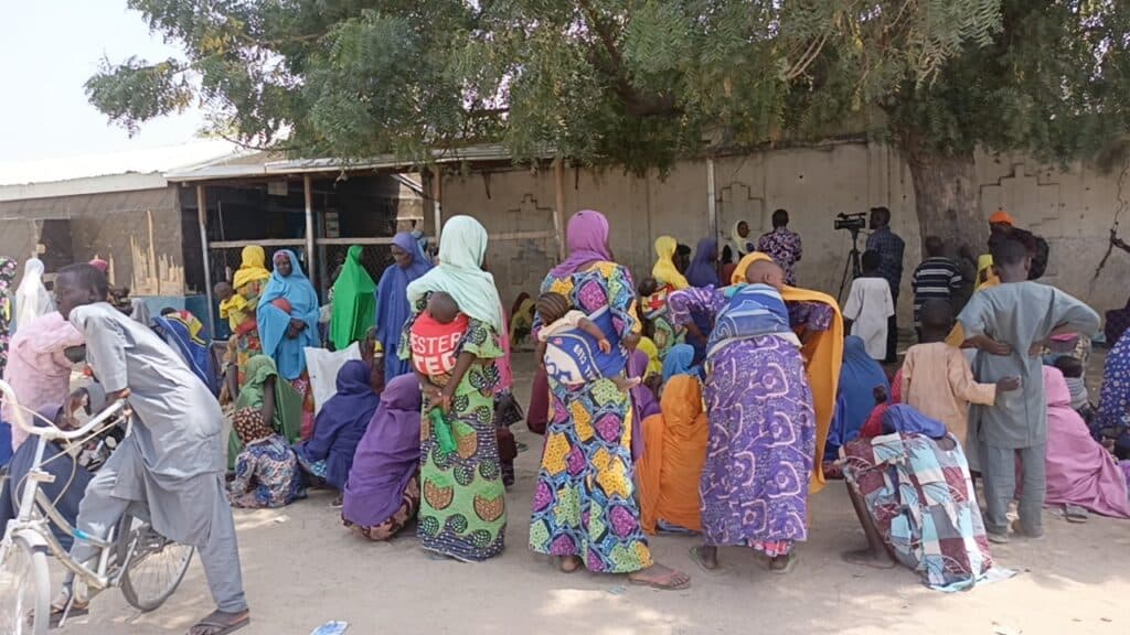 A group of people in colorful clothing gathering at a rural community center under trees.