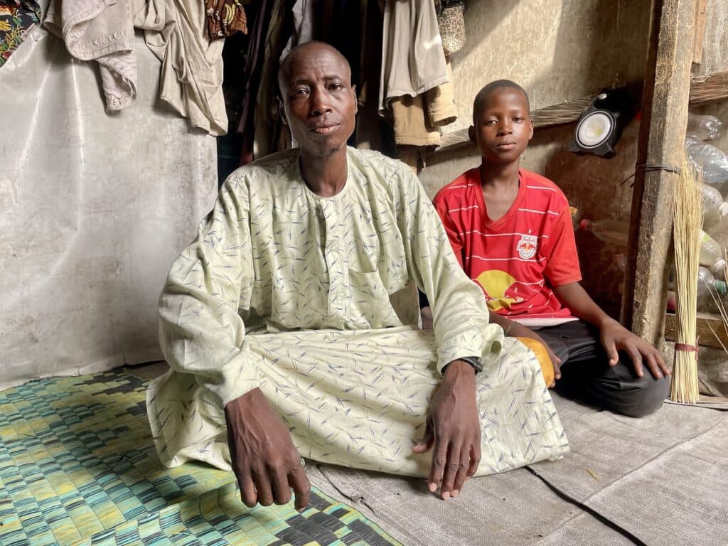 An adult male in traditional attire and a young boy in a red jersey sitting on a mat inside a room.