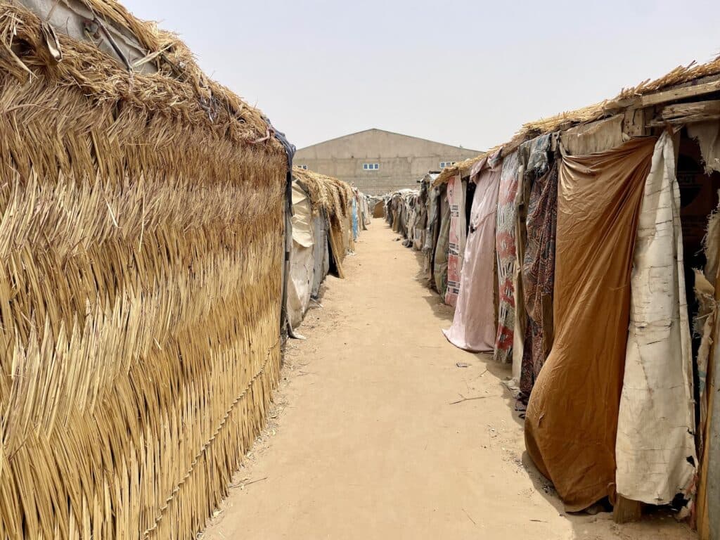 A sandy pathway flanked by makeshift shelters with thatched and tarp-covered walls in a makeshift settlement.