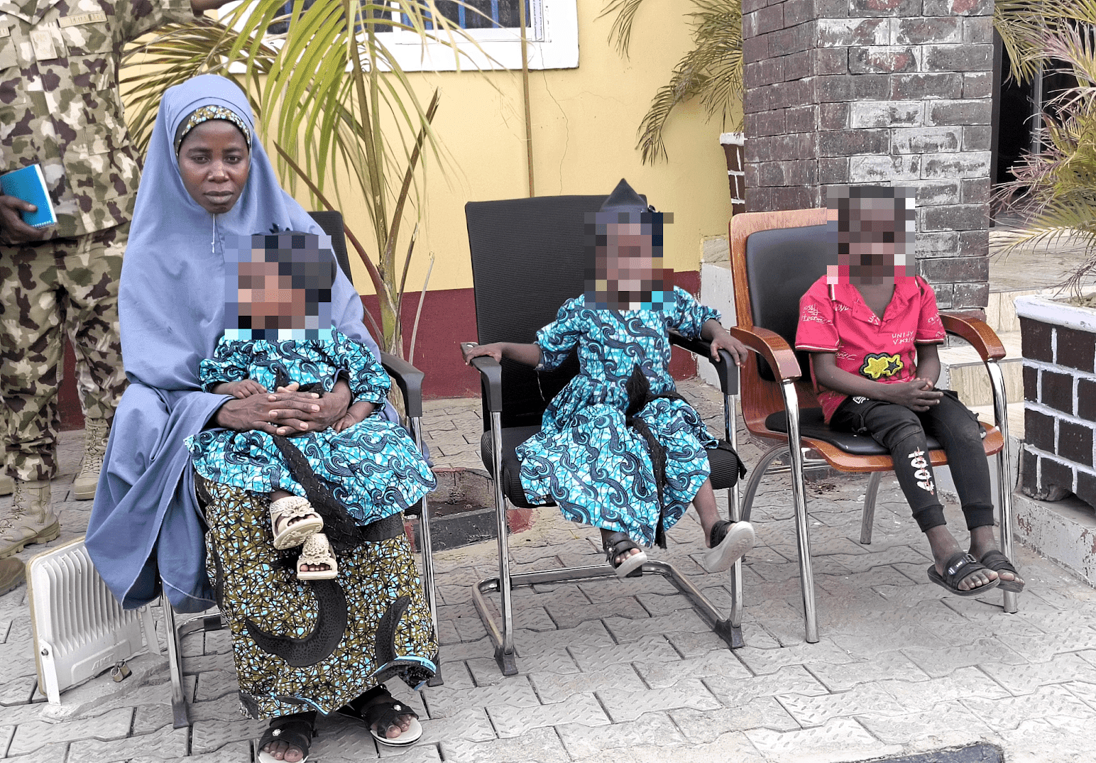 Woman in blue hijab sitting next to children in matching outfits, faces pixelated for privacy.