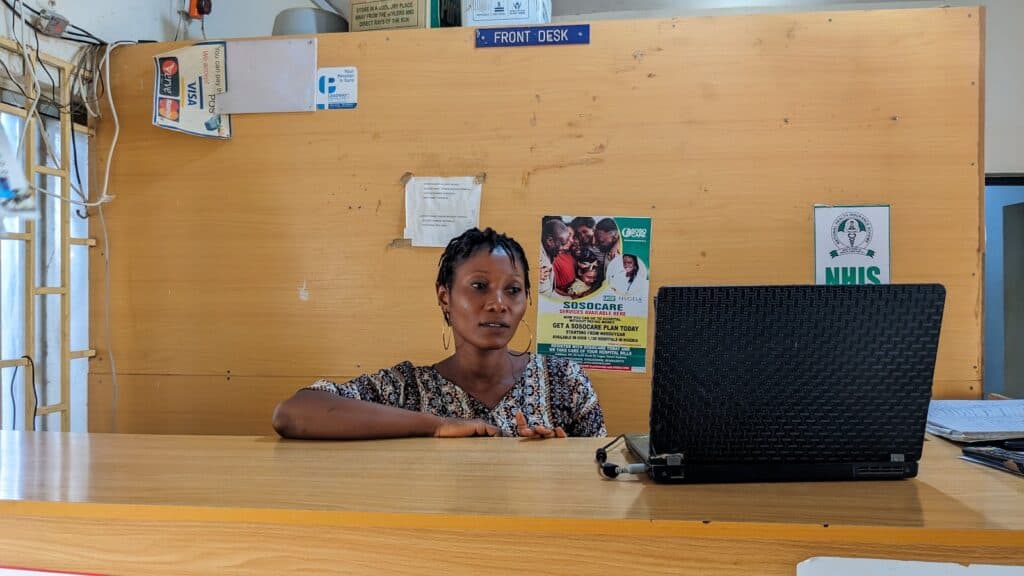 Woman sitting at a front desk with a laptop and posters in the background.