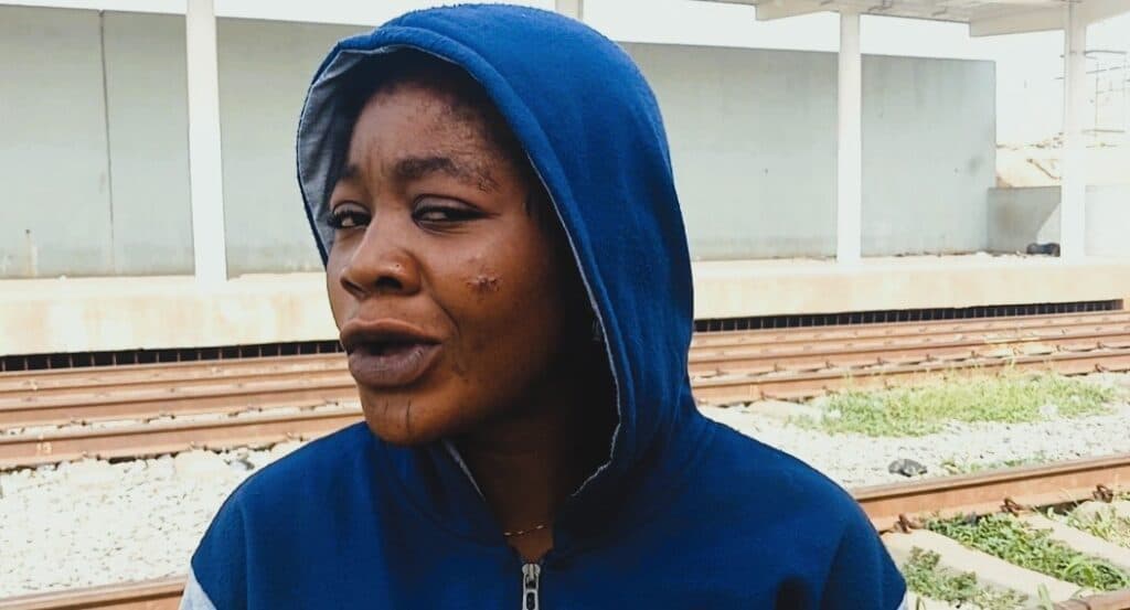 A person wearing a blue hoodie stands near train tracks, appearing contemplative or concerned.