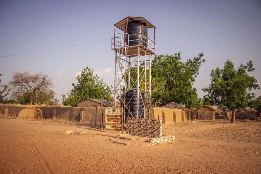 Water tower with a metal tank on a structure amid a rural landscape with huts and trees under a blue sky.