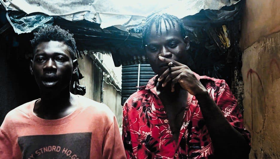 Two men standing in a narrow alleyway, one smoking, with a makeshift tarp overhead.