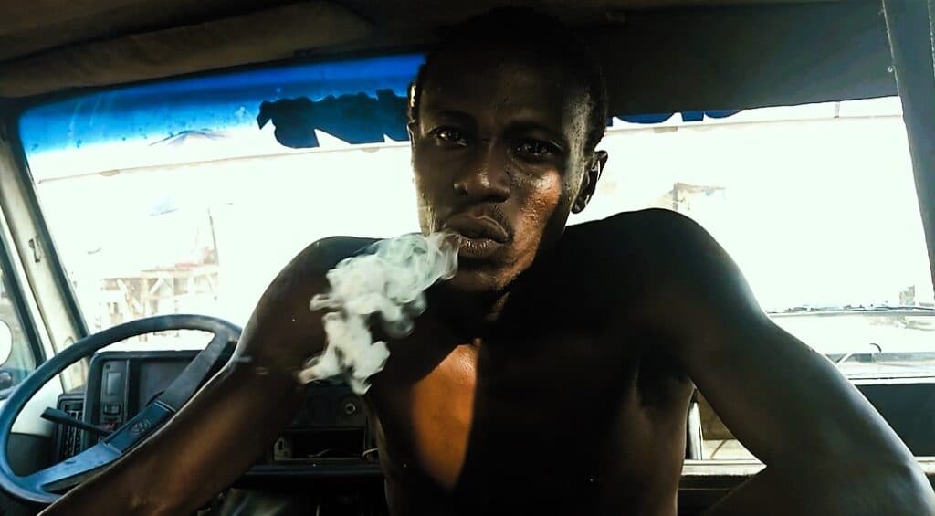 Man exhaling smoke while sitting inside a vehicle with sunlight streaming in.