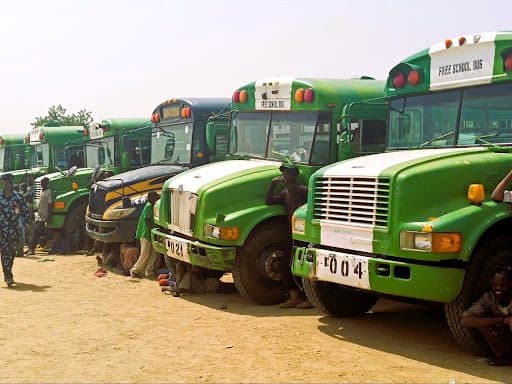 A row of green and white school buses parked with people standing and sitting nearby.