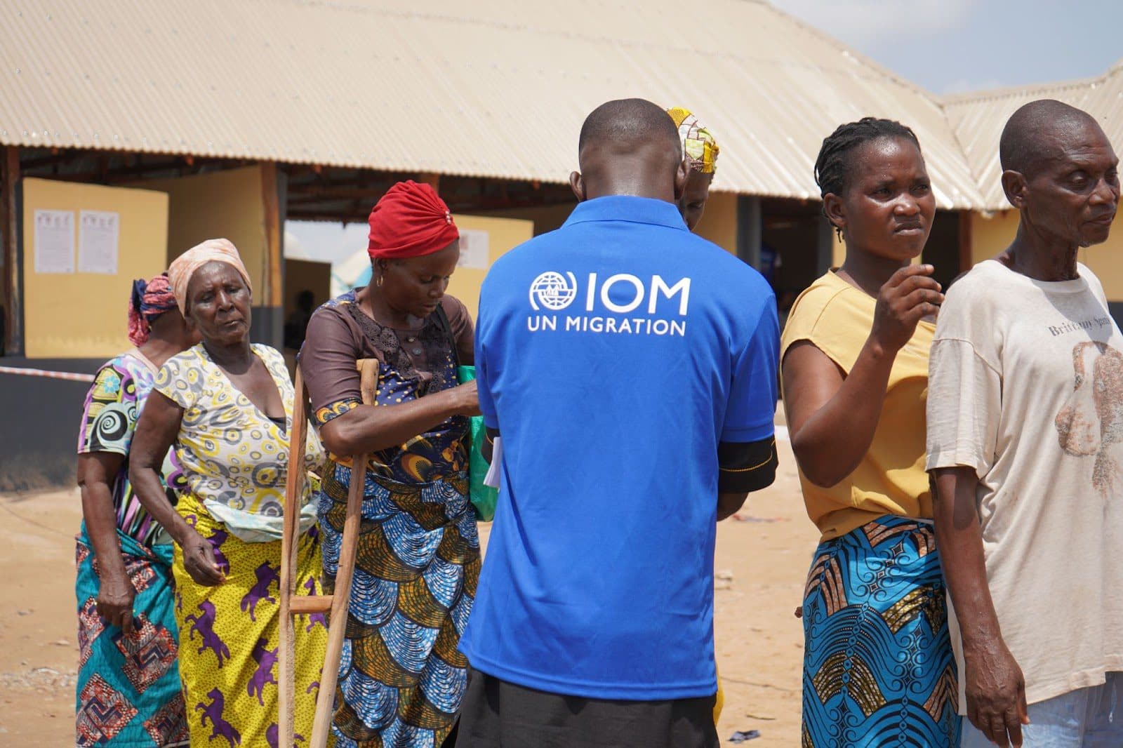 IOM staff member in blue shirt with locals in a sunny outdoor setting, likely during a migration-related activity.