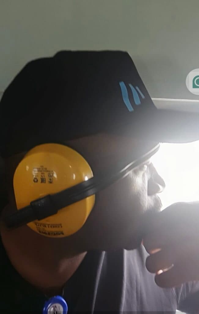 Person wearing a cap and yellow ear defenders, looking pensive, hand on chin.