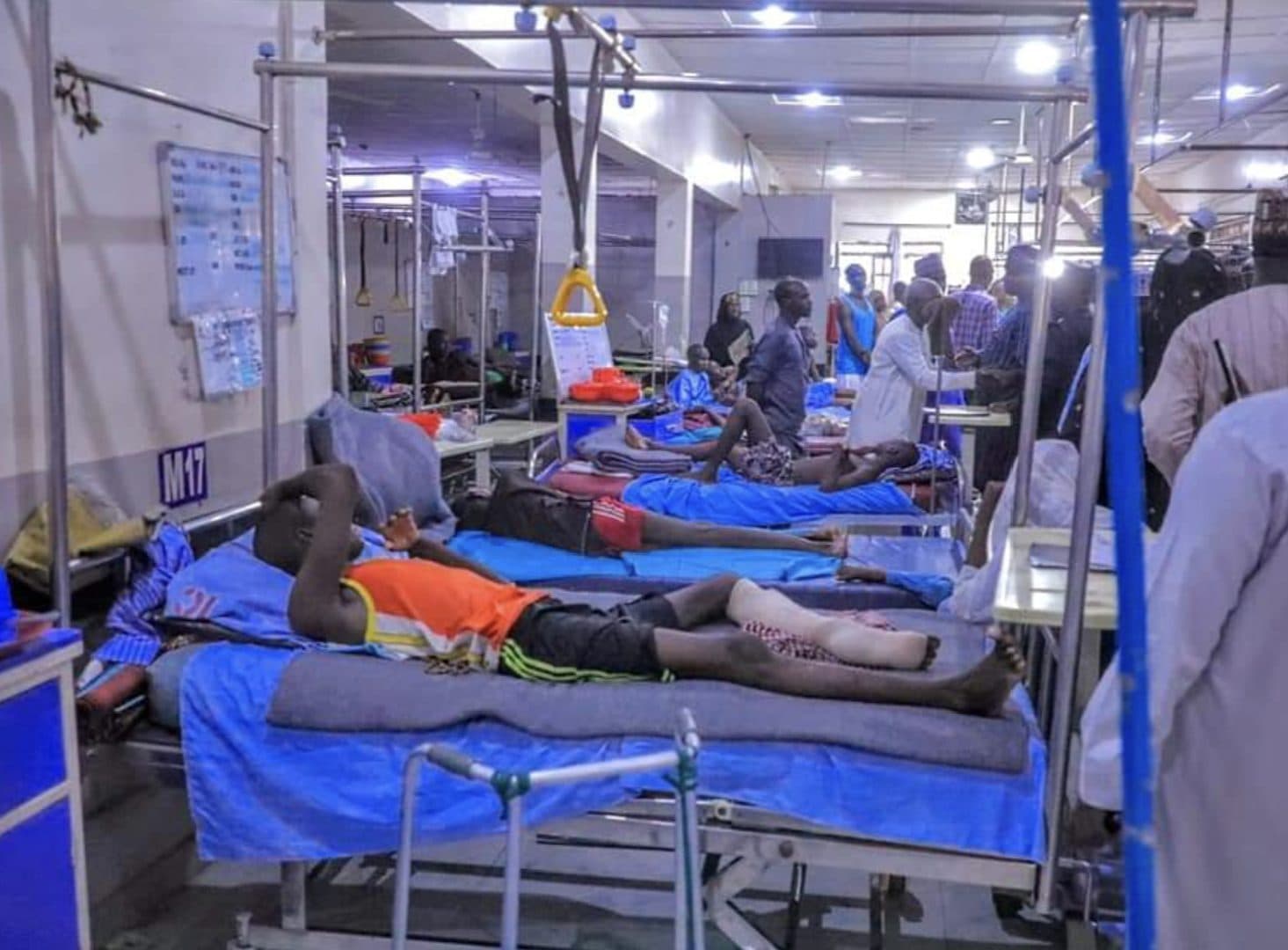 A crowded hospital ward with patients on beds and people walking between them.