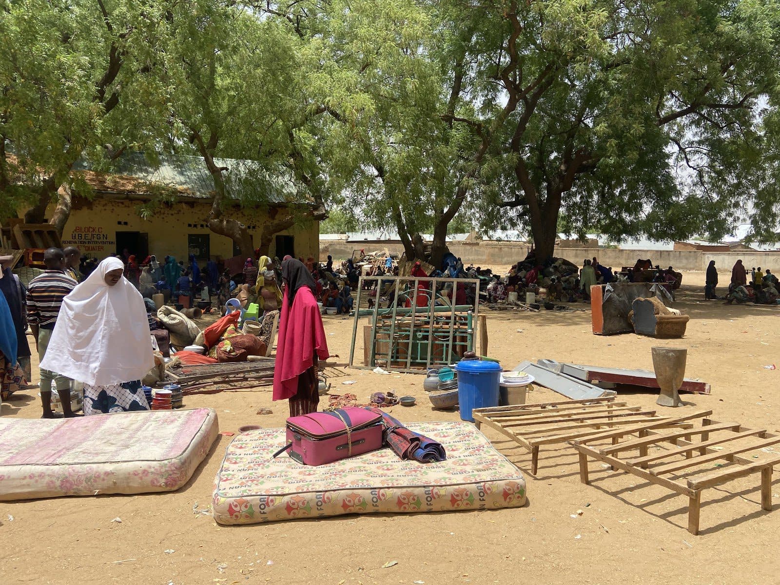 A bustling outdoor marketplace under trees with people and goods, including mattresses and containers.
