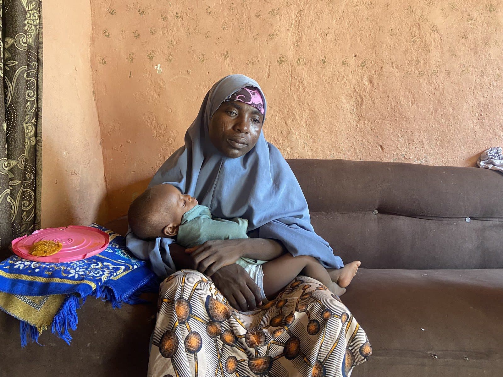 Woman in a headscarf holding a sleeping child on a sofa, with a colorful plate beside her.