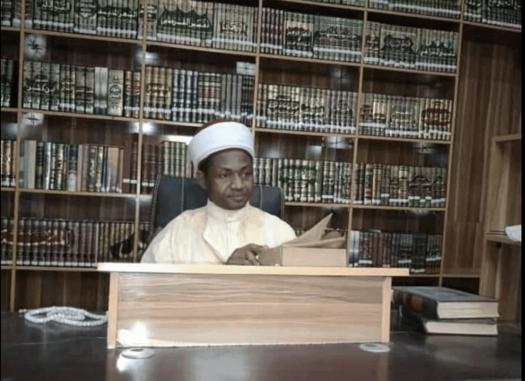 Man in a traditional cap reading a book at a desk with a large bookshelf filled with books behind him.