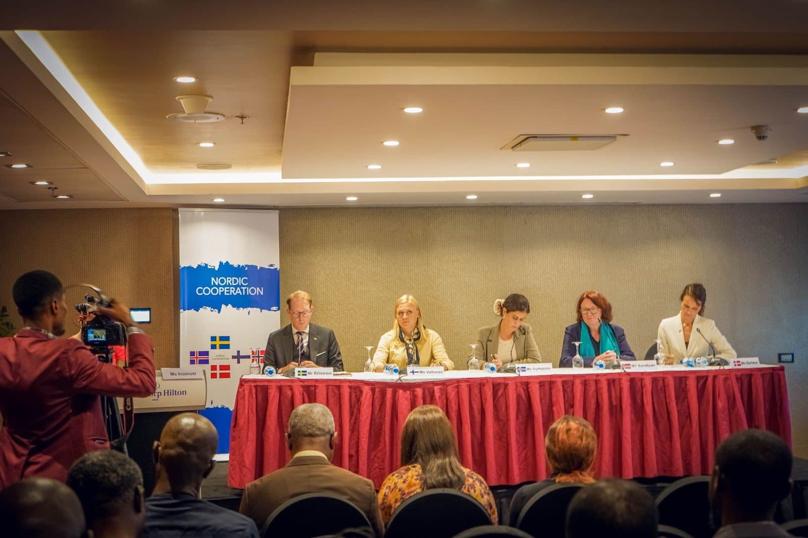 Panel of five individuals at a Nordic Cooperation conference with an audience and a cameraman in the foreground.
