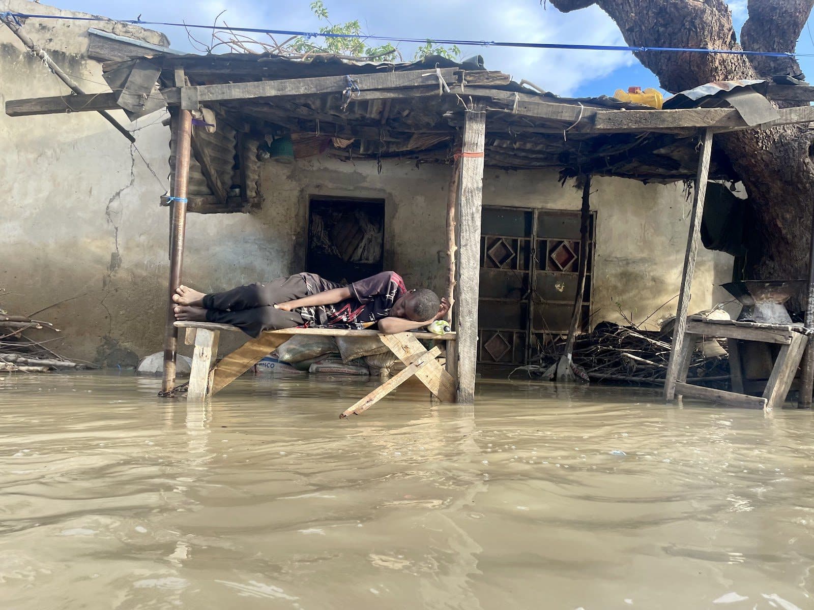 Person sleeping on makeshift bed above floodwaters near damaged house.