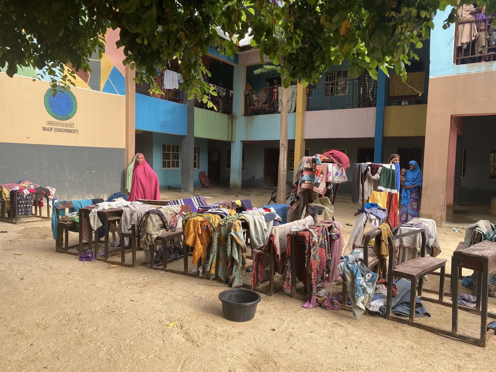 Colorful clothing laid out on tables and benches to dry in a sunlit courtyard, with women standing nearby.
