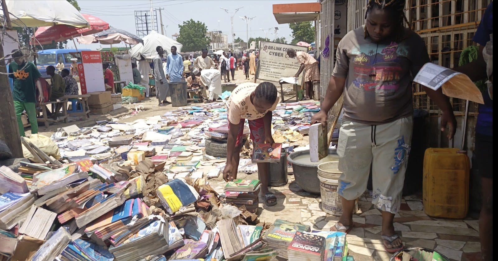 Street book market with people browsing piles of books on the ground under the sun.