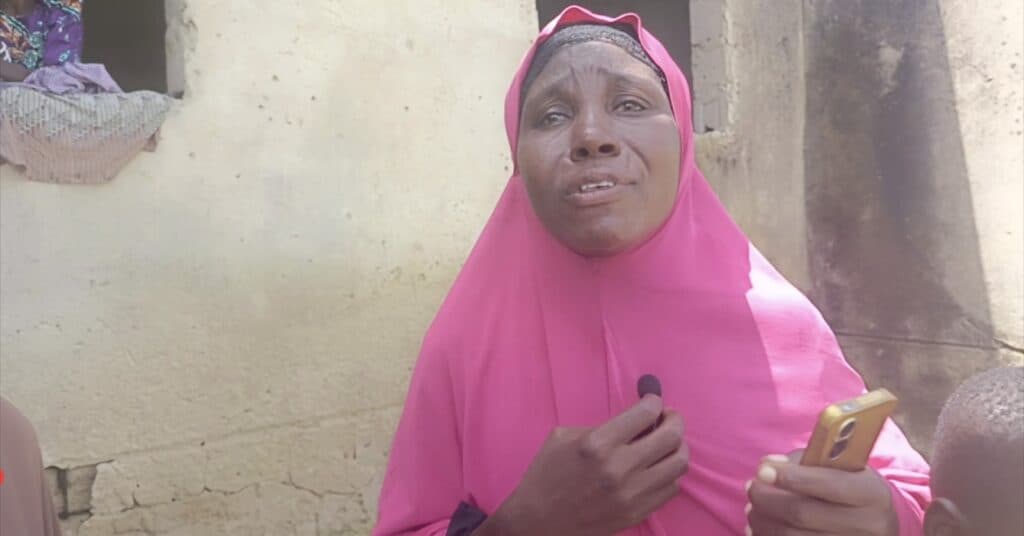 Woman in pink hijab speaking, holding a microphone, with a smartphone in her other hand, standing against a weathered wall.