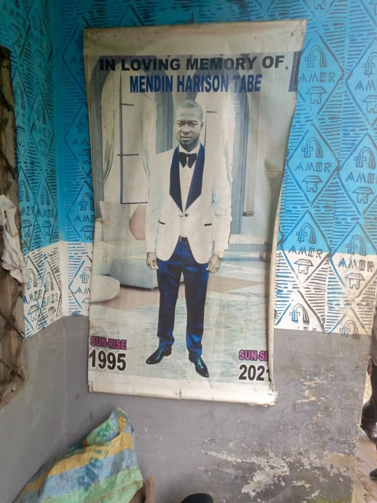 A memorial poster of a man in a suit, with text "In Loving Memory" and the years 1995 - 2021, displayed against a patterned wall.