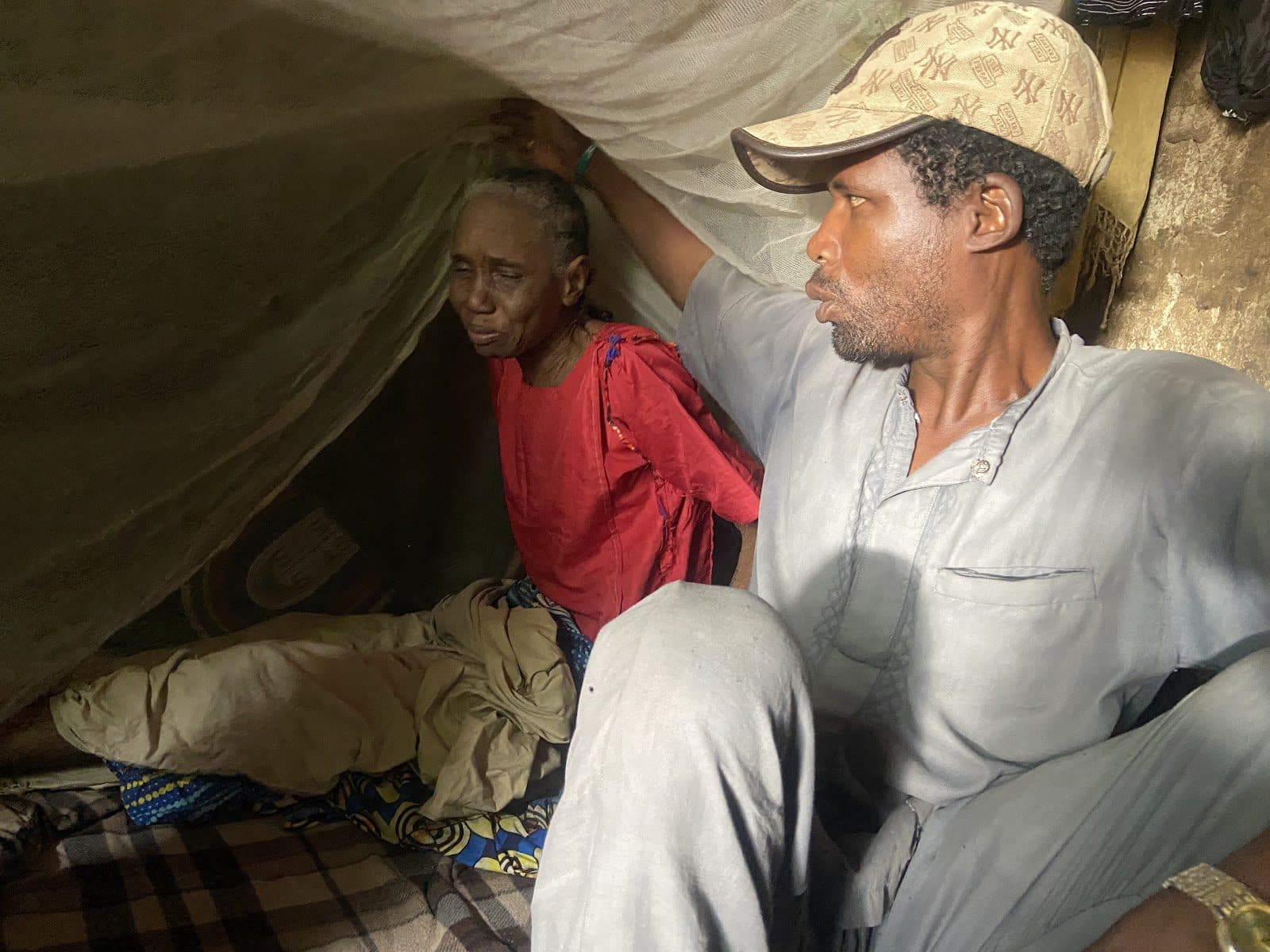 Two people sitting under a makeshift tent, one looking weary and the other gazing into the distance.
