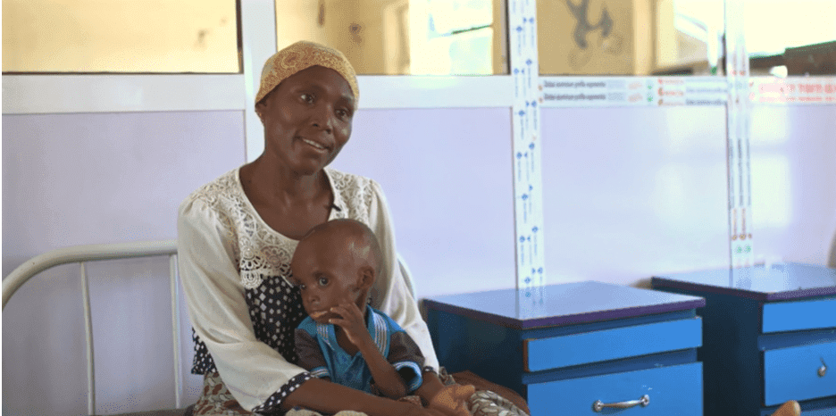Woman in a headscarf sitting with small child on her lap, indoors near hospital beds.