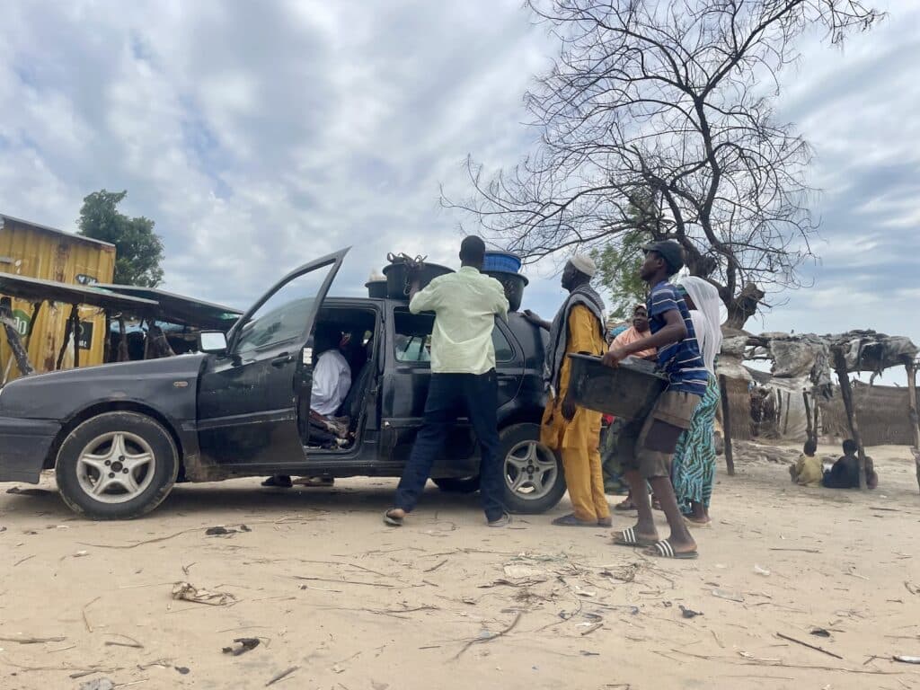 People loading items into a car in a sandy area with makeshift structures and a leafless tree under a cloudy sky.