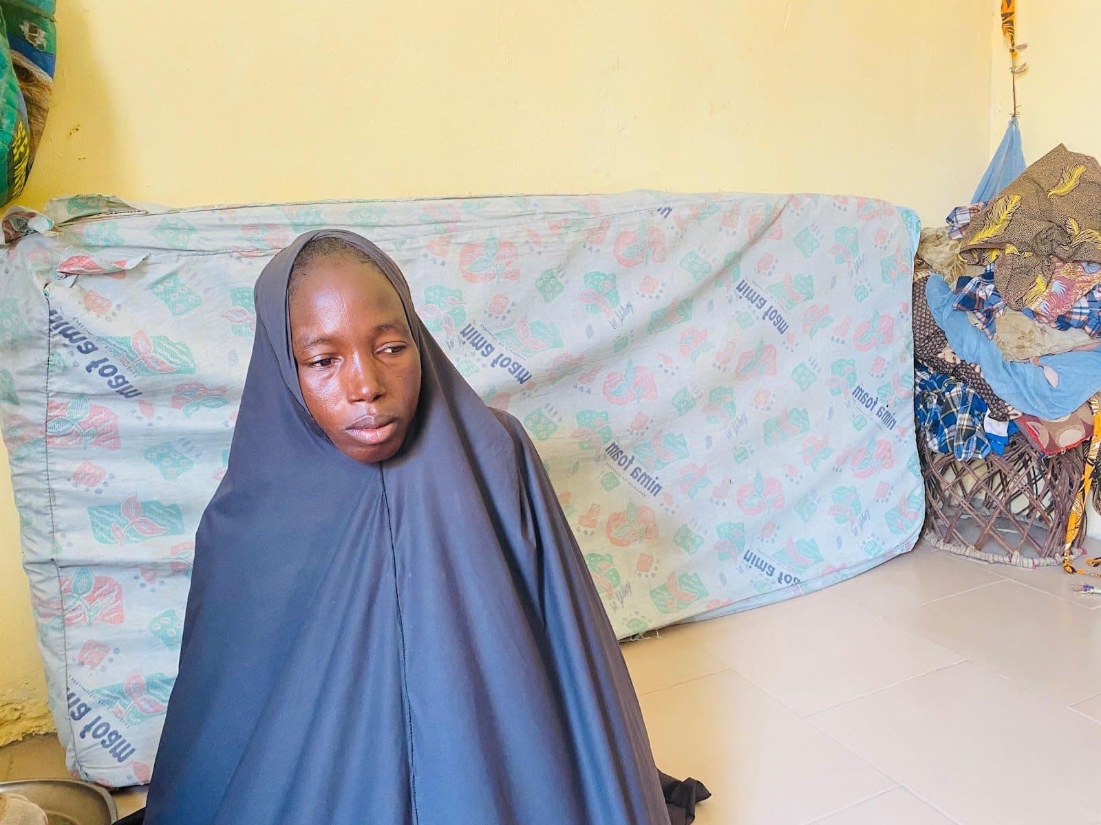 Person in a dark hijab sitting in front of a cloth-draped background in a room.