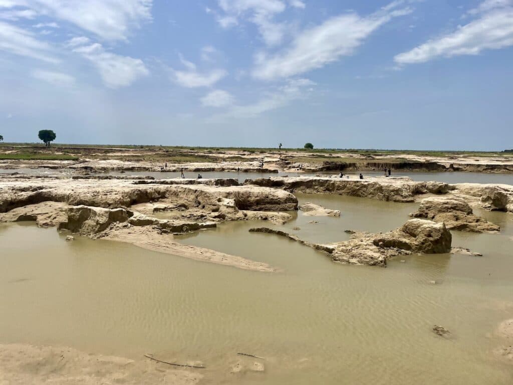 Sandy landscape with water pools under a cloudy sky, with sparse vegetation and distant figures.