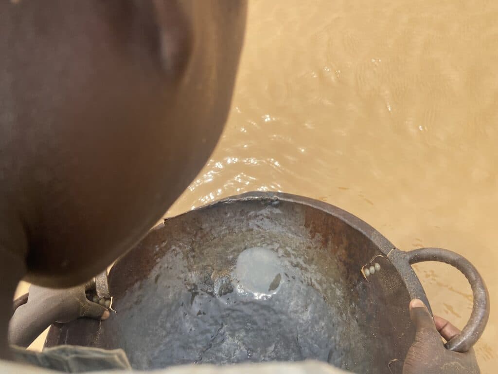 Over-the-shoulder view of a person holding a pan near murky water, possibly panning for minerals.