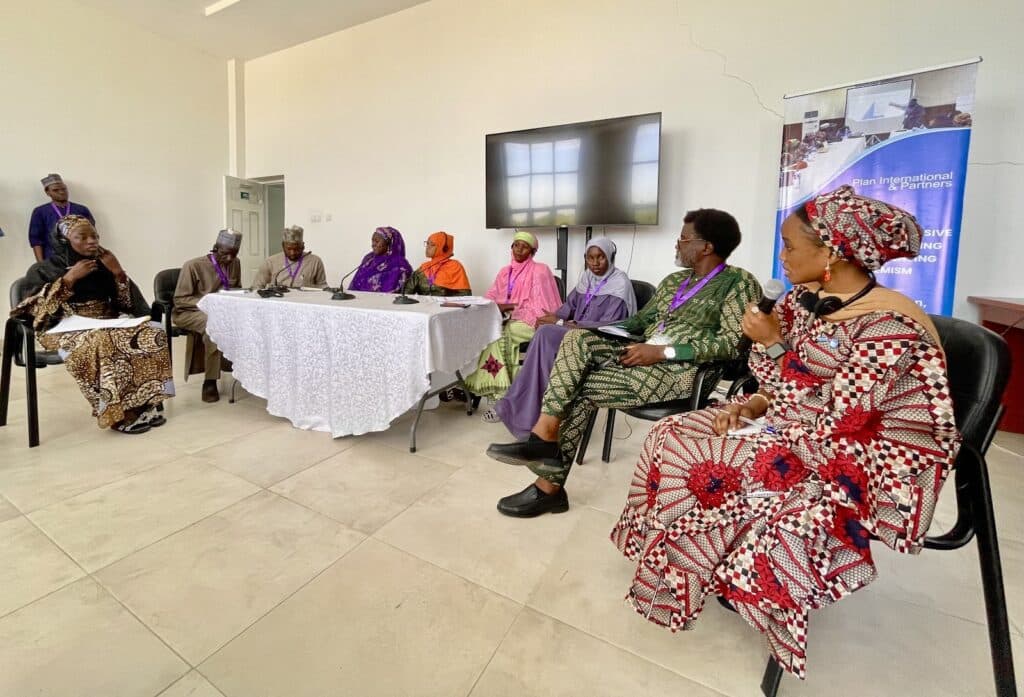 A group of people, wearing colorful attire, seated in a panel discussion setup with microphones and a banner saying "Plan International & Partners."
