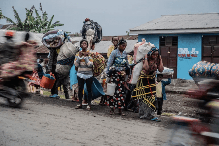 People carrying belongings walk along a road, with a blurred motorcycle passing by.