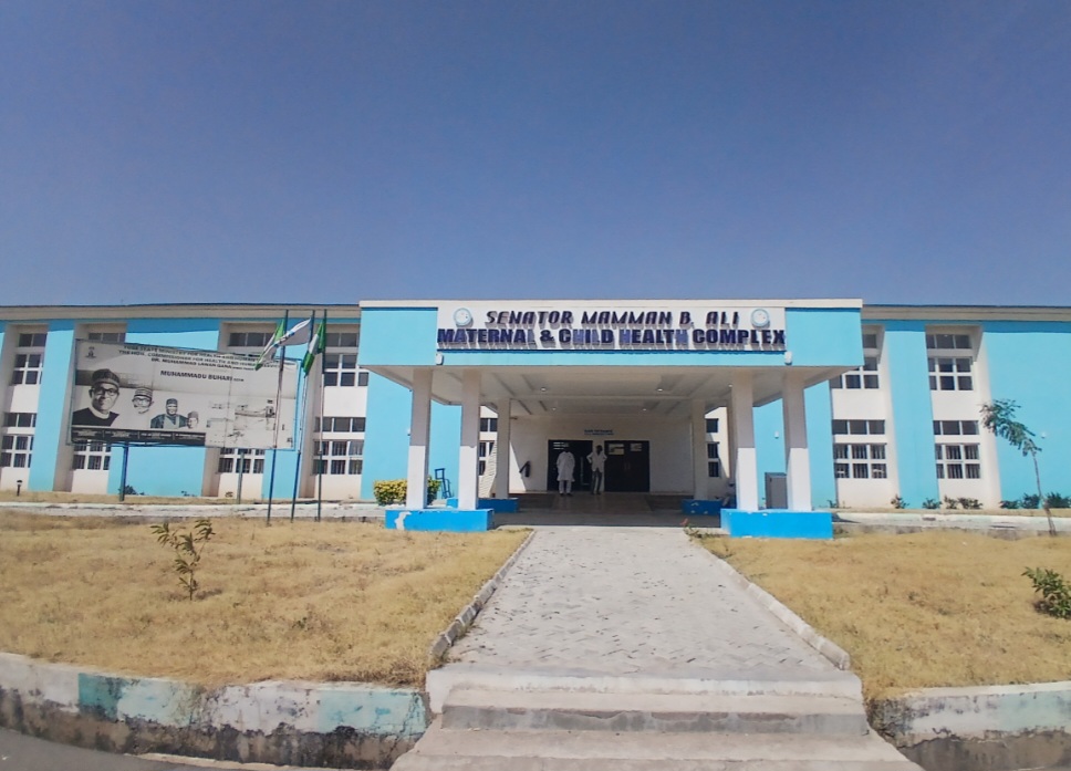 Blue and white building of the Senator Mamman B. Ali Maternal & Child Health Complex, with a billboard and flags in the foreground.