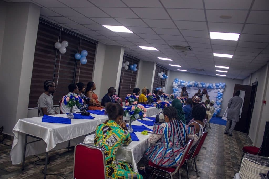 People seated at a decorated banquet hall with blue and white balloons, listening to speakers at the front.