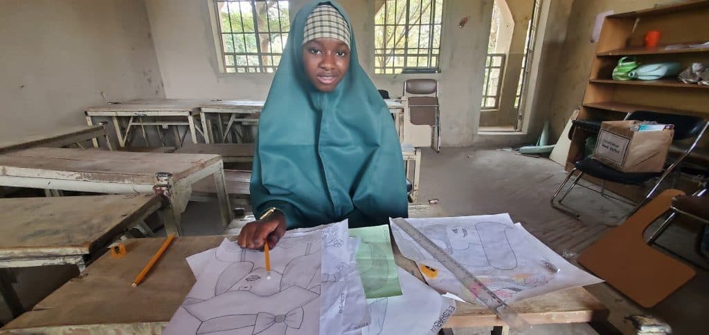 Young girl in a classroom, wearing a green hijab, seated at a desk drawing with pencils and papers scattered around.