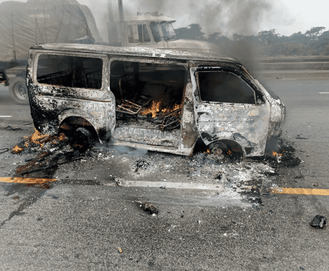 Burned-out van with flames inside, on a highway. Smoke rises, and a truck is visible in the background.
