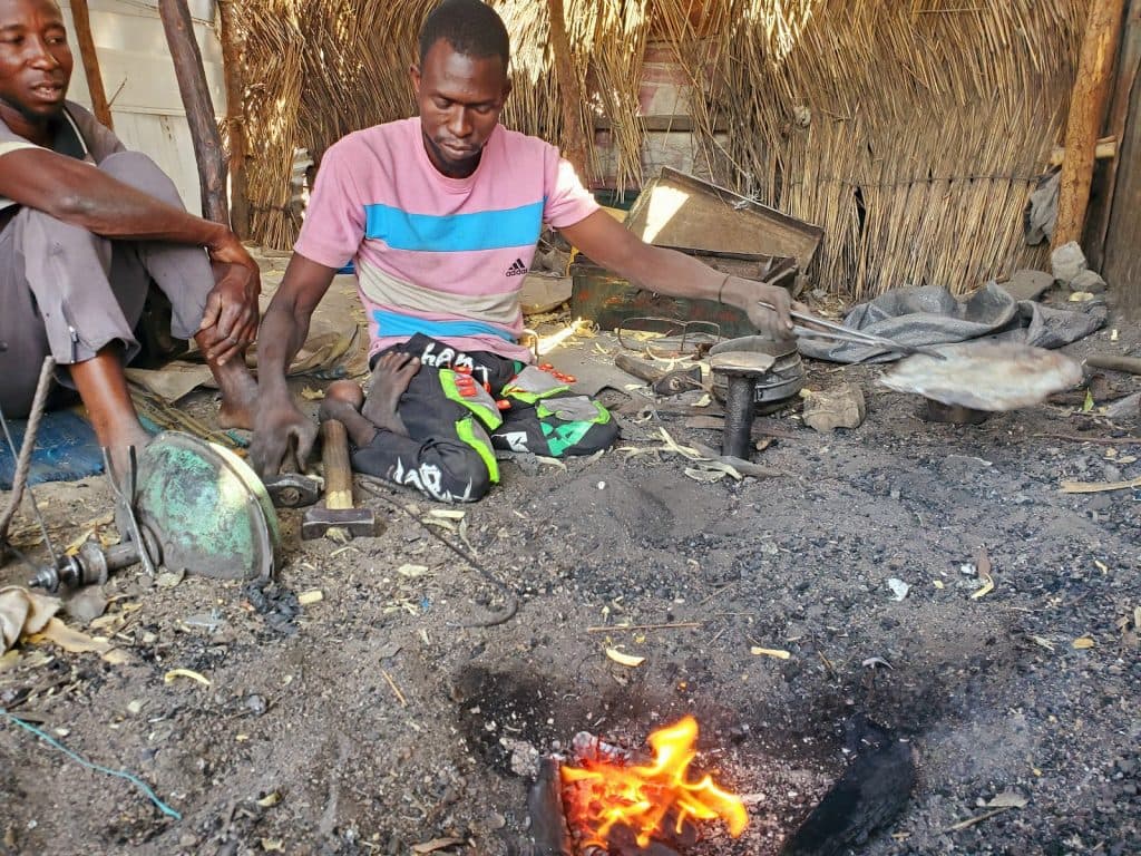 A man works with tools and a small fire in an outdoor setting, while another person sits nearby.