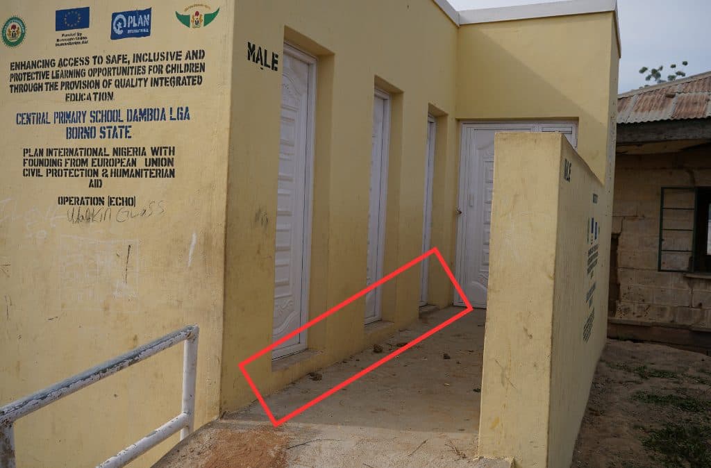 Yellow building with white doors labeled "Male." Text on wall about educational access. Ramp with railing. Borno State, Nigeria.