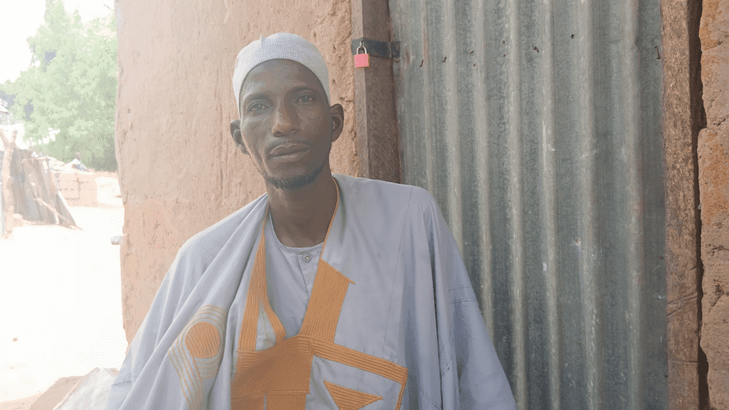Man in traditional attire stands outside near a corrugated metal wall and a mud building, looking at the camera.