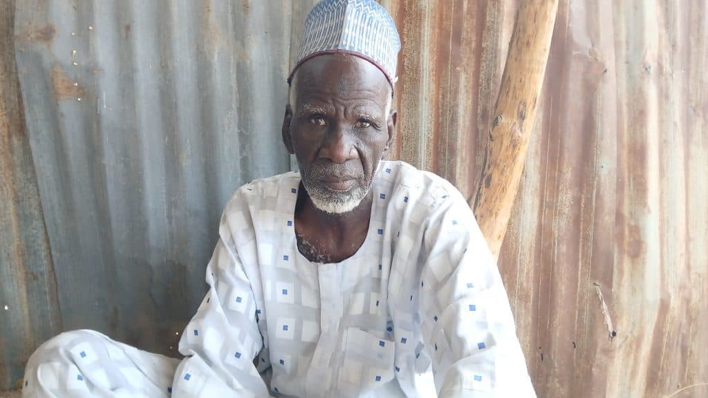 Elderly man in traditional attire, sitting against a corrugated metal wall, looking forward.