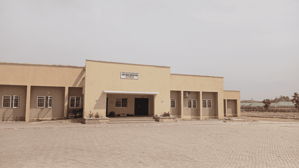 Beige building with a sign "Cottage Hospital Gombi." Paved area in front, windows on the side, and a motorcycle parked nearby.