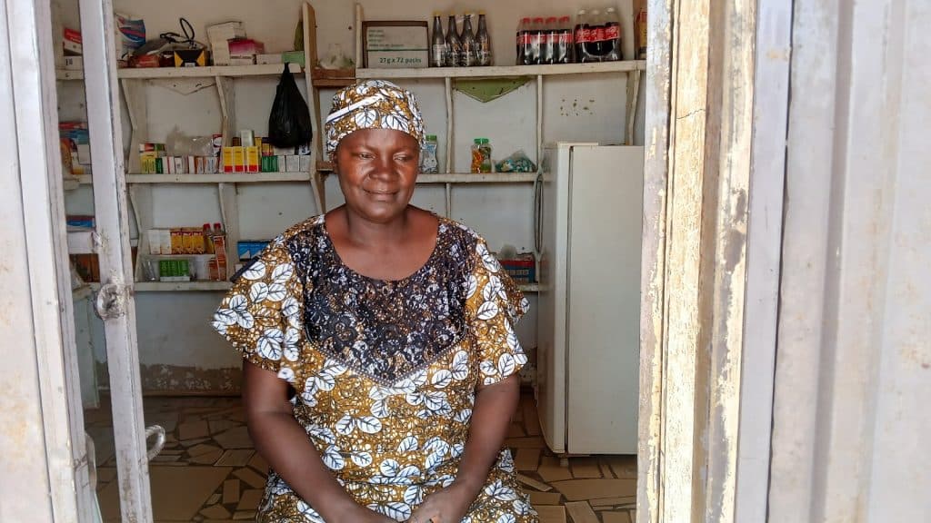Woman in patterned dress and headscarf sitting in a small shop with shelves stocked with items and beverages.